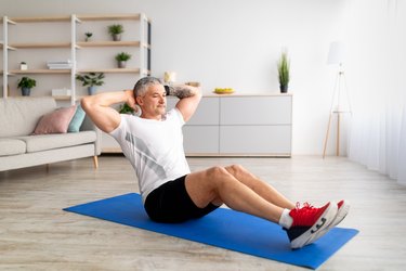 Older adult in workout clothes doing a sit-up on an exercise mat in their living room to demonstrate a body-weight workout for older adults