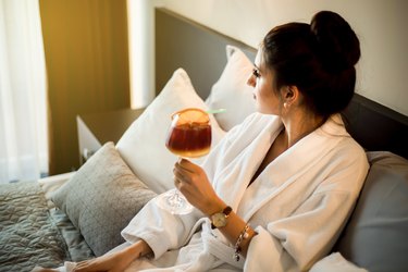 A young woman in a bathrobe enjoys bed and drinks a cocktail